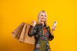 © deagreez - Glamorous mature woman with shopping bags smiling while using smartphone against yellow background