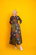 © deagreez - Mature blonde woman in colorful patchwork dress stands with hands on hips smiles against bright yellow background, promoting fashion and lifestyle