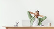 © Prostock-studio - A young man relaxes during a break at his desk. He leans back in his chair, stretching his arms and smiling. There is a laptop, a notebook, and stationery on the desk.