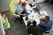 © DragonImages - Three middle aged men including Caucasian foreman discussing construction project at office desk, reviewing blueprints and digital plans on laptop, safety vest and helmet visible nearby