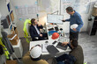 © DragonImages - Group of middle aged Caucasian men and one Black man discussing construction blueprints in office, foreman standing and pointing at monitor while others sitting with laptops and hard hats