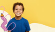 © Prostock-studio - A black boy stands happily against a yellow background. He wears a blue shirt and has curly hair. His smile shows joy and youthfulness. This scene captures a lively moment in time.