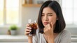 © Rakibul - Woman experiencing tooth pain while drinking a cold beverage in a bright kitchen.
