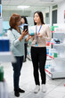 © DC Studio - Woman shows her smartphone screen to pharmacist, standing in store aisle filled with pharmaceutical products. Customer presents product QR code on phone to drugstore attendant inside chemist shop