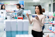 © DC Studio - Calm Asian female customer stands in pharmacy aisle comparing medicine packaging and product labels. Modern healthcare retail environment represents informed decision making and wellness shopping