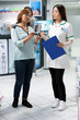 © DC Studio - Customer speaks with drugstore attendant about promotional package shown on flyer. Friendly pharmacist holding clipboard explains suitable pharmaceutical products to client in pharmacy aisle.