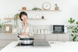 © buritora - A young Asian woman in an apron cooking in front of an induction stove in the kitchen, thinking and worrying