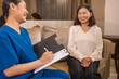 © Nalinee - A professional Asian nurse in blue scrubs holding a clipboard, discussing health results or a treatment plan with a senior Asian woman sitting on a sofa during a home care visit.