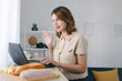 © Aphisara - Young woman smiling and waving during an online video call while working at her desk, illustrating remote work, virtual communication, home office lifestyle, and modern digital interaction.