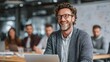 © Prostock‑studio - Successful bearded man with glasses happily smiles at the camera in a modern brightly lit office environment working on his laptop with professional colleagues
