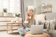 © Pixel-Shot - Young woman drinking coffee while using laptop on floor in cozy bright living room with flowers, remote work, online learning and relaxed home lifestyle