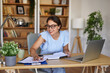 © Stockphotodirectors - Woman in a blue blouse sits at a wooden desk, smiling while writing in a notebook, surrounded by plants and books in a bright and inviting home office space, highlighting focused productivity.