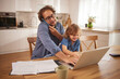 © Stockphotodirectors - A father talks on the phone while his young child plays on a laptop, showing a moment of family interaction and shared activities at home.