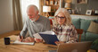 © Stockphotodirectors - An older couple reviews life insurance documents at home. One partner writes notes while the other looks through a folder. A laptop and a coffee cup rest on the table.