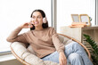 © Pixel-Shot - Beautiful young woman in headphones listening to audiobook and sitting on armchair near window at home