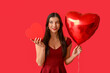 © Pixel-Shot - Beautiful young woman with kiss marks on her face holding gift box and heart shaped air balloon on red background