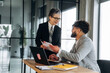 © standret - Business professionals in a modern office discussing project details, with a laptop and documents on a wooden table, large windows providing natural light