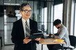 © standret - Mature businesswoman in formal attire holding a tablet smiles while standing in a modern office with a colleague working at a desk in the background