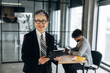 © standret - Senior businesswoman in formal attire holding a tablet smiles confidently in a modern office with a colleague working at a desk in the background