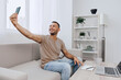 © SHOTPRIME STUDIO - Smiling man taking a selfie at home, showcasing a cozy living room atmosphere with modern decor, capturing a moment of happiness and connection.