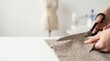 © 24DIY - Close up of hands cutting textured fabric with black scissors on a white table Blurred tailoring equipment behind