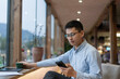 © zhu difeng - A middle-aged Asian man wearing glasses uses his smartphone while sitting by the window in a cozy café with mountain view
