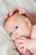 © Daria - Close up portrait of baby with parent hand on head in soft natural light, infant self soothing with fingers in mouth, protection and parental care concept