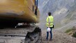 © DawDunia - Female engineer holding hard hat and tablet, supervising excavator on a rugged mountain construction site, overseeing civil engineering work and project progress outdoors