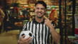 © Krakenimages.com - Young hispanic man dressed as a soccer referee holding a ball smiles confidently while gesturing in a retail clothing store, showcasing sports and leisure in an indoor setting.