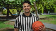 © Krakenimages.com - Young man in referee uniform smiling and holding basketball in a green outdoor park setting.