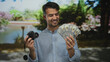 © Krakenimages.com - Young hispanic man holding camera and displaying u.s. dollars in a lush green park setting, conveying success and leisure.