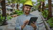 © Krakenimages.com - Young hispanic man with hardhat holding tablet in green outdoor park setting, smiling confidently amidst palm trees and modern buildings suggesting urban development innovation