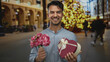 © Krakenimages.com - Young man holding bouquet and heart-shaped gift on city street with festive lights in background, smiling cheerfully.