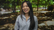 © Krakenimages.com - Young chinese woman smiling in a green park with trees in the background, wearing a striped shirt, exuding happiness and joy in an outdoor setting.