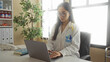 © Krakenimages.com - Young chinese woman doctor in uniform using a laptop in a clinic room, holding her shoulder with a stethoscope draped around her neck, conveying a sense of workplace strain.