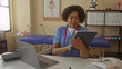 © Krakenimages.com - Woman in clinic wearing scrubs and stethoscope holds tablet and types on laptop at desk with shelf of binders and anatomy poster behind; focus.