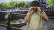 © Krakenimages.com - Young hispanic man rubbing his hands over his eyes on a street bridge by a canal, beige shirt and wristbands visible, boat and crowd behind; fatigue solitude.