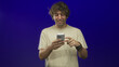 © Krakenimages.com - Man tapping smartphone with finger in studio blue backdrop, wearing beige tshirt and bracelets, smiling while interacting with the screen; playful curiosity.