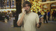 © Krakenimages.com - Man holding creditcard and talking on phone in building decorated for holidays, wearing glasses and bracelets, inspecting cards near lit christmas tree and crowds; payment worry.