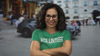 © Krakenimages.com - Middle age hispanic woman volunteer wearing green shirt crosses arms and smiles in bustling street square; confidence support commitment.