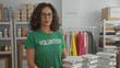 © Krakenimages.com - Hispanic woman volunteer in a green uniform stands confidently indoors in a charity room surrounded by donation items and packaged food.
