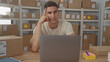 © Krakenimages.com - Young man with finger to temple working on laptop amid stacked parcels on shelves in delivery building; concentration.