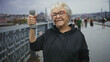 © Krakenimages.com - Senior woman smiling holds a gray dumbbell in raised right hand on street beside metal railing, wearing hoodie and colorful glasses; confidence wellness.