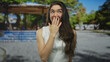 © Krakenimages.com - Young hispanic woman in white tank top yawning with hand covering mouth in sunny green park setting; surprise.