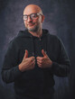 © mark_gusev - A man wearing a black hoodie and glasses is smiling. He is standing in front of a grey background. Classic portrait of a confident and friendly looking male model.