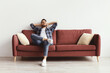 © Prostock-studio - A man sits back on a red couch in a bright room. He has a big smile on his face and hands behind his head. The space is light and features two cushions.