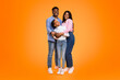 © Prostock-studio - A family stands closely together with smiles on their faces. They are dressed casually. The background is bright orange. The scene shows warmth and happiness in their expressions.