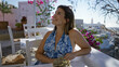 © Krakenimages.com - Woman smiling with bare shoulder visible, seated at a terrace restaurant building in santorini wearing sunglasses and a blue sundress; vacation serenity.