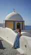© Krakenimages.com - Woman leaning on a white wall beside a greek church building in santorini, shielding her eyes and looking up toward the sky; serenity travel.