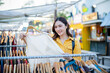 © Laingzaifa - An Asian woman is happily browsing for fashionable clothes in a boutique, smiling as she shops, holding shopping bags, showcasing the enjoyment of modern shopping.